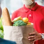 Asian deliver man wearing face mask in red uniform handling bag of food, fruit, vegetable give to female costumer in front of the house. Postman and express grocery delivery service during covid19.