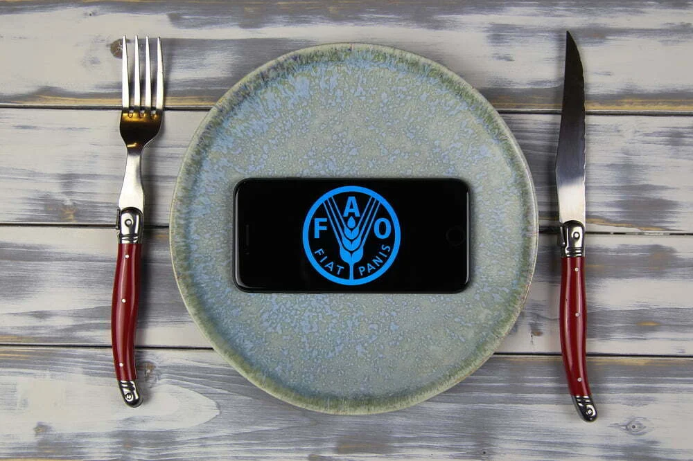 Prato com smartphone ao centro exibindo o logótipo da FAO, acompanhado por talheres, simbolizando tecnologia e alimentação global