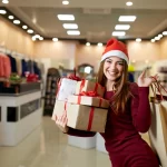 Happy girl shopping gifts in mall on christmas sale. New year holidays shopping idea concept. Smiling woman with colorful paper presents bags and gift boxes wearing christmas hat in store or shop.