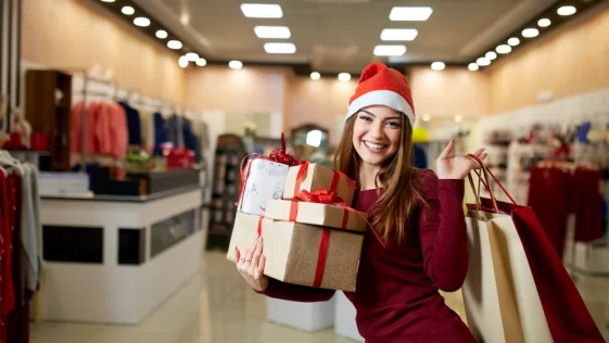 Happy girl shopping gifts in mall on christmas sale. New year holidays shopping idea concept. Smiling woman with colorful paper presents bags and gift boxes wearing christmas hat in store or shop.