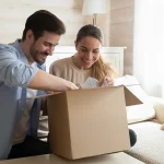 Happy young couple unpacking awaited parcel at home together, curious man and woman looking into open cardboard box, satisfied customers received online store order, good delivery service