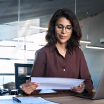 Focused latin hispanic young business woman working on laptop computer reading financial document report in office. Accountant entrepreneur manager businesswoman doing paperwork using pc. Copy space