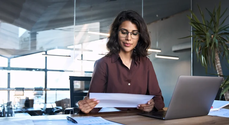 Focused latin hispanic young business woman working on laptop computer reading financial document report in office. Accountant entrepreneur manager businesswoman doing paperwork using pc. Copy space