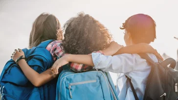 Rear view of multiethnic elementary middle school kids pupils classmates friends with backpack in casual clothes are hugging embracing each other standing outdoors.