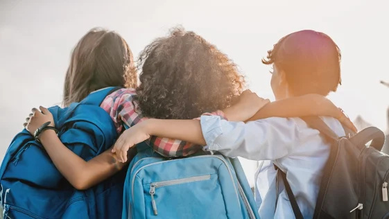 Rear view of multiethnic elementary middle school kids pupils classmates friends with backpack in casual clothes are hugging embracing each other standing outdoors.