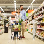conceito de venda, consumismo e pessoas - família feliz com criança e carrinho de compras comprando comida em mercearia ou supermercado