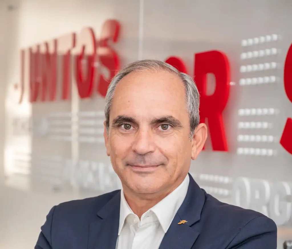 Retrato de um homem de fato e camisa branca, em frente a um fundo com letras vermelhas desfocadas