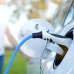 Electric car or ev is charging at station . man use the white power cable and plug on nature background. eco and clean energy concept with the mobile phone smart technology. zero gas no transmission