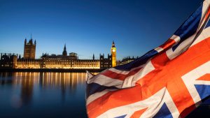 Bandeira do Reino Unido em primeiro plano, com o Palácio de Westminster e o Big Ben iluminados junto ao rio Tamisa