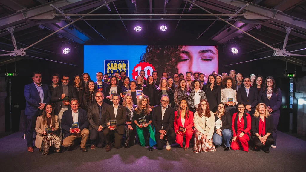 Fotografia de grupo dos vencedores e participantes na cerimónia Sabor do Ano 2026, em palco, com prémios na mão e ecrã oficial ao fundo