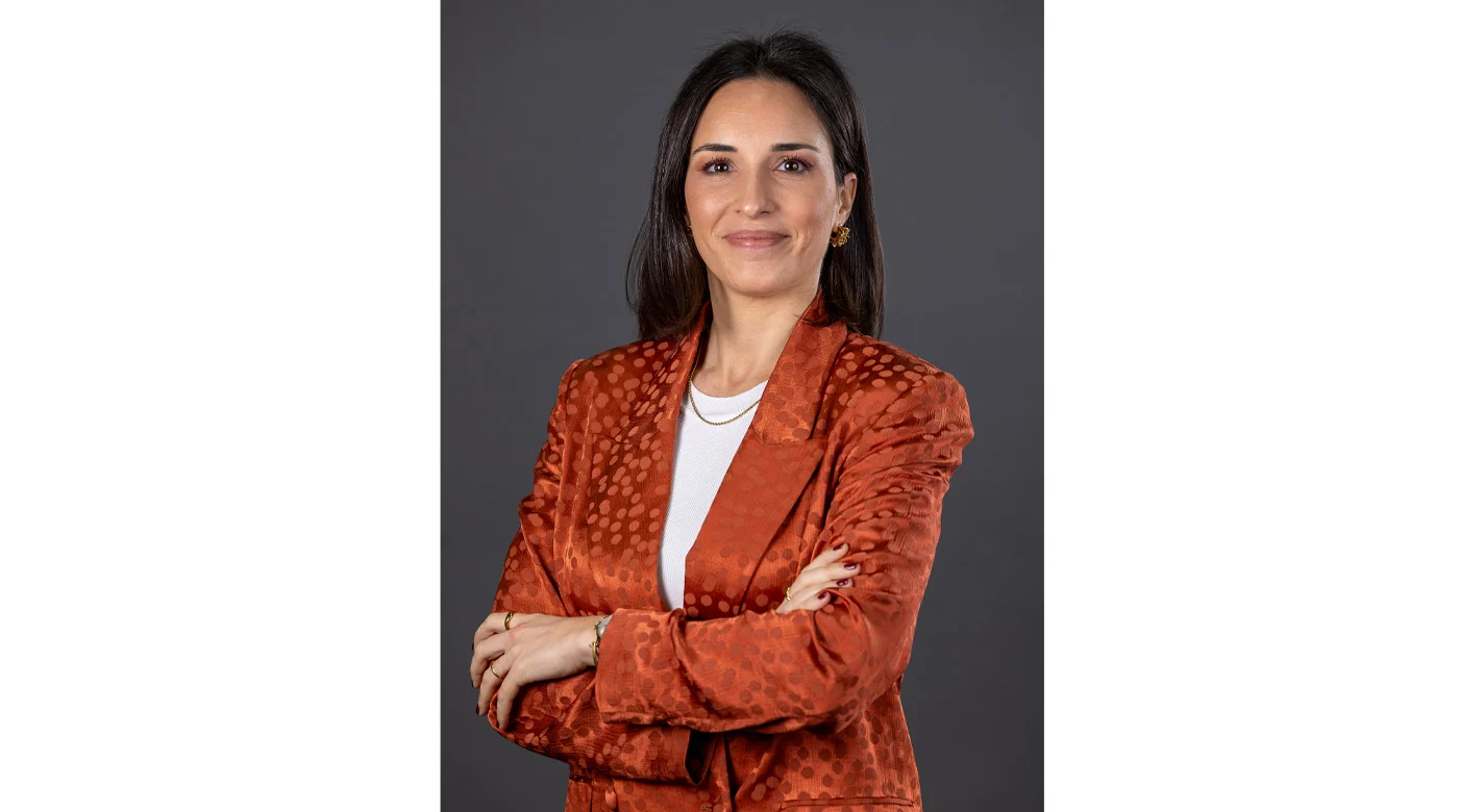 Retrato de mulher em ambiente de estúdio, com blazer laranja e braços cruzados, sobre fundo neutro