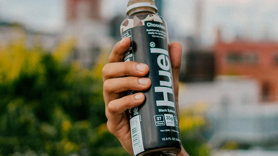 Mão a segurar uma garrafa de bebida nutricional Huel sabor chocolate, em ambiente exterior desfocado