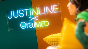 Letreiro luminoso com as marcas “Just In Line x OralMED”, em ambiente moderno e colorido, com uma pessoa desfocada em primeiro plano