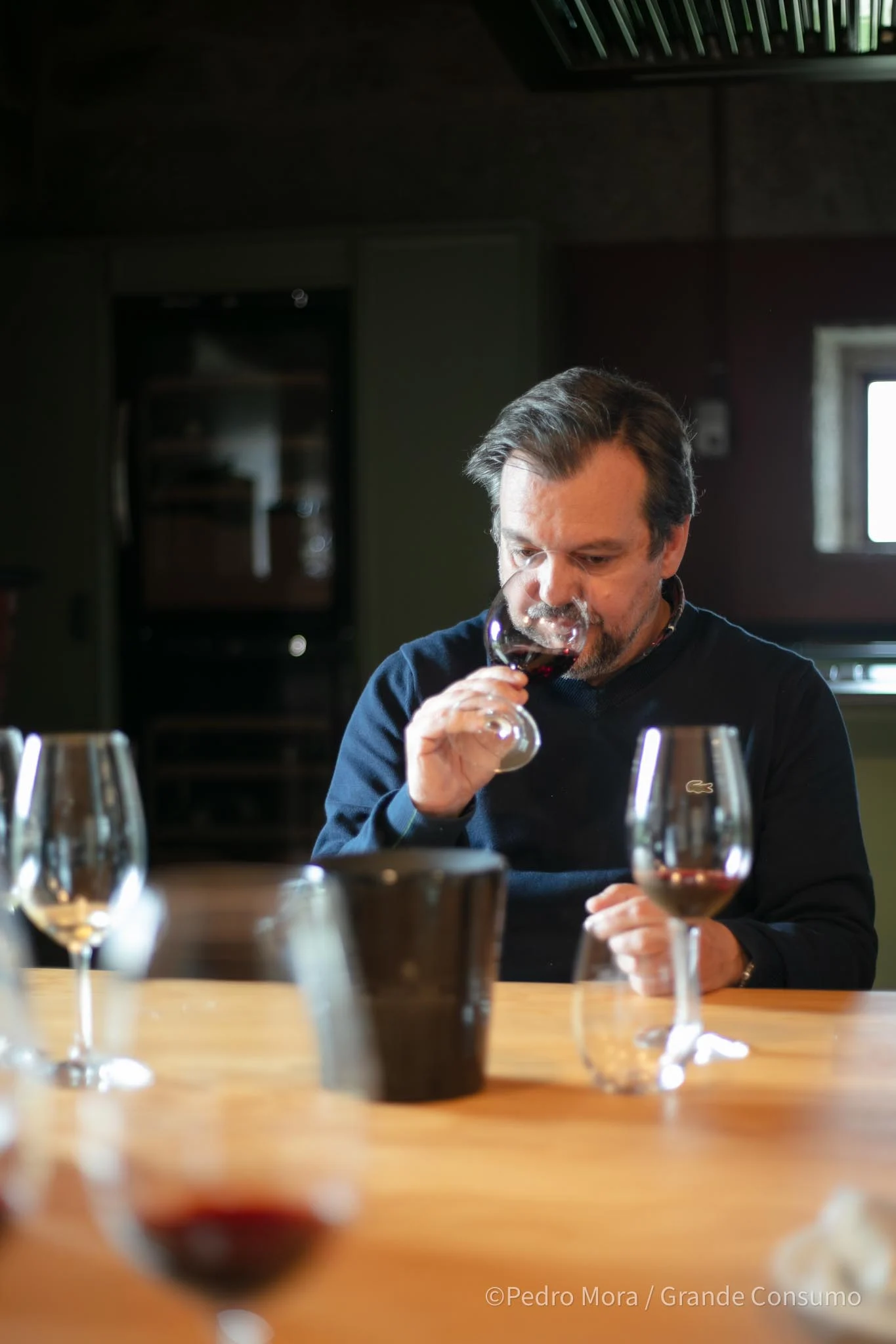 Homem cheira um copo de vinho numa mesa rodeada por copos de vinho