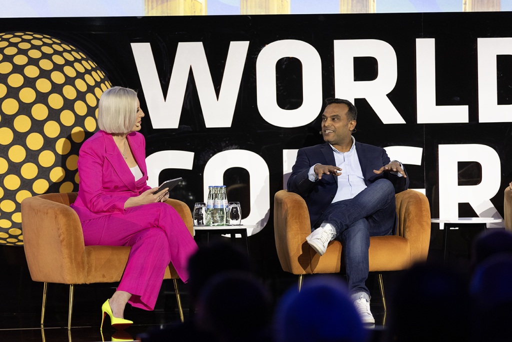 Imagem de um painel no World Retail Congress, com dois oradores em palco: uma moderadora vestida de rosa e um convidado em traje informal elegante, sentados em poltronas e em conversa. Ao fundo, destaca-se o branding do evento