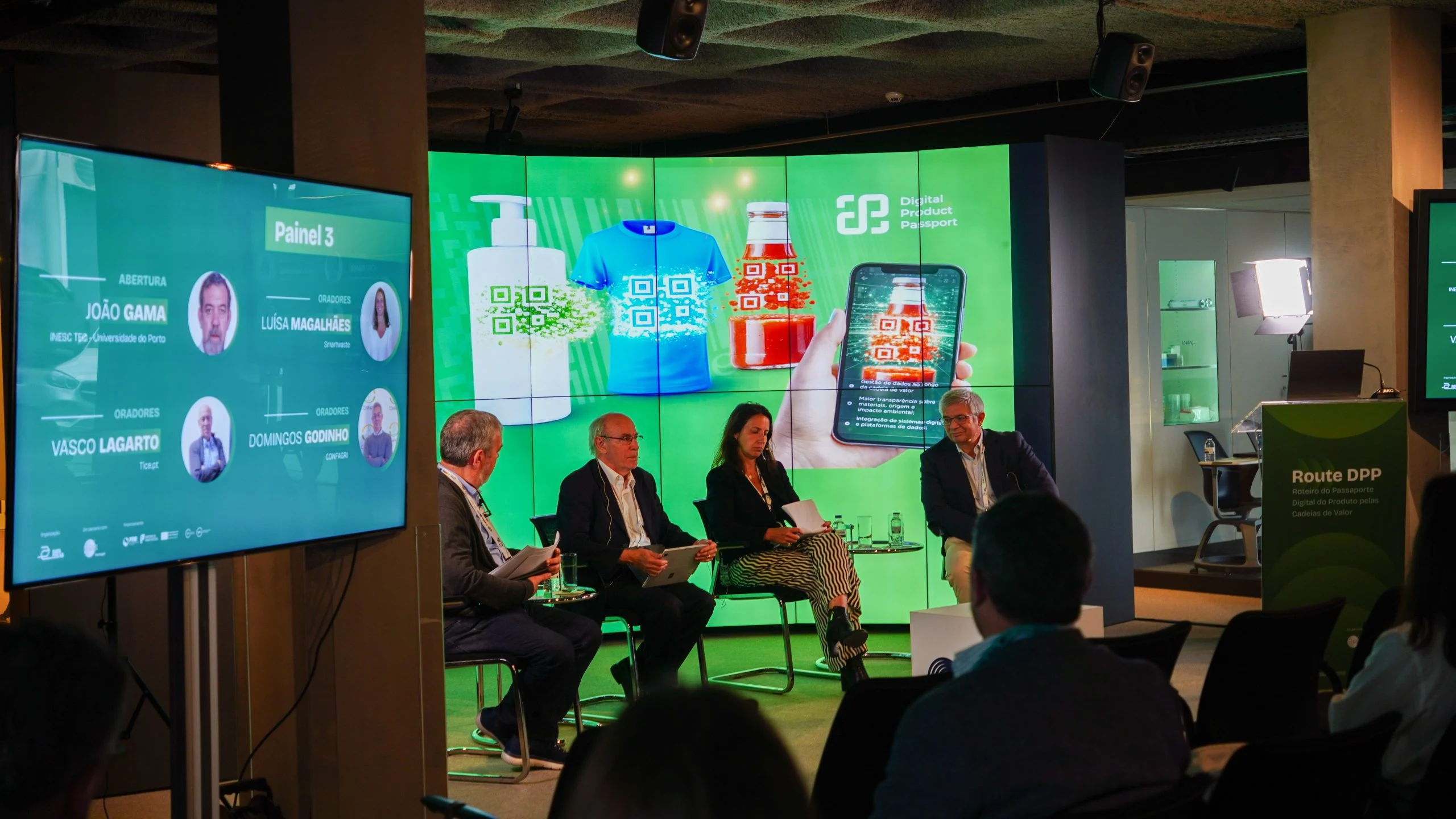 Painel de debate sobre o Passaporte Digital do Produto num evento dedicado às cadeias de valor e sustentabilidade. Vários oradores estão sentados em palco perante um ecrã gigante com exemplos de produtos e códigos QR associados ao conceito de Digital Product Passport, enquanto a audiência acompanha a sessão