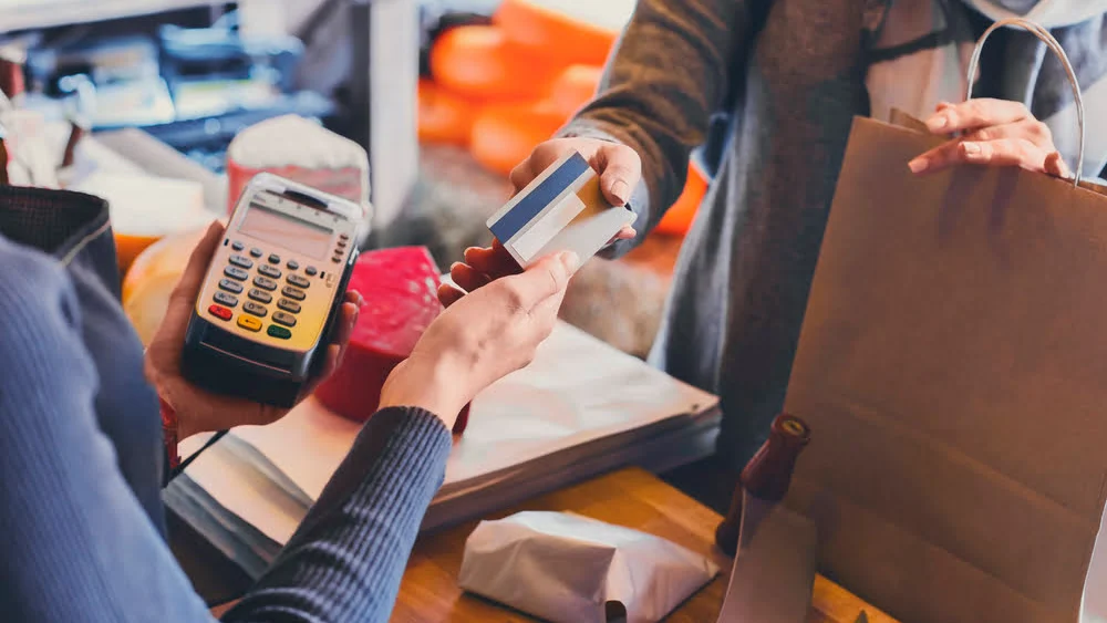 Cliente efetua pagamento com cartão num ponto de venda, enquanto segura um saco de compras, ilustrando uma experiência de compra em ambiente de retalho