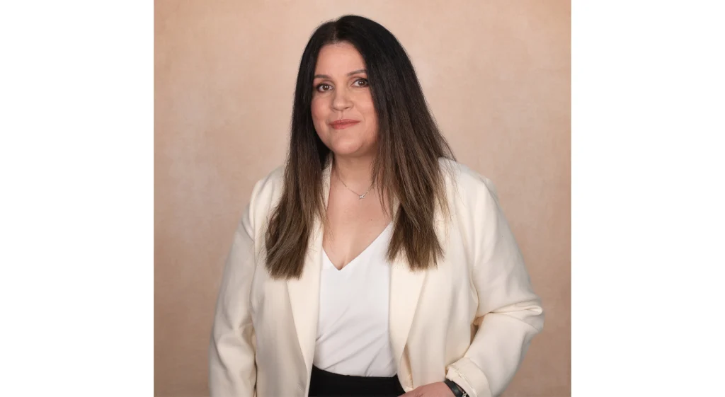 Mulher de cabelo comprido escuro, com blazer claro, em pose de estúdio sobre fundo bege
