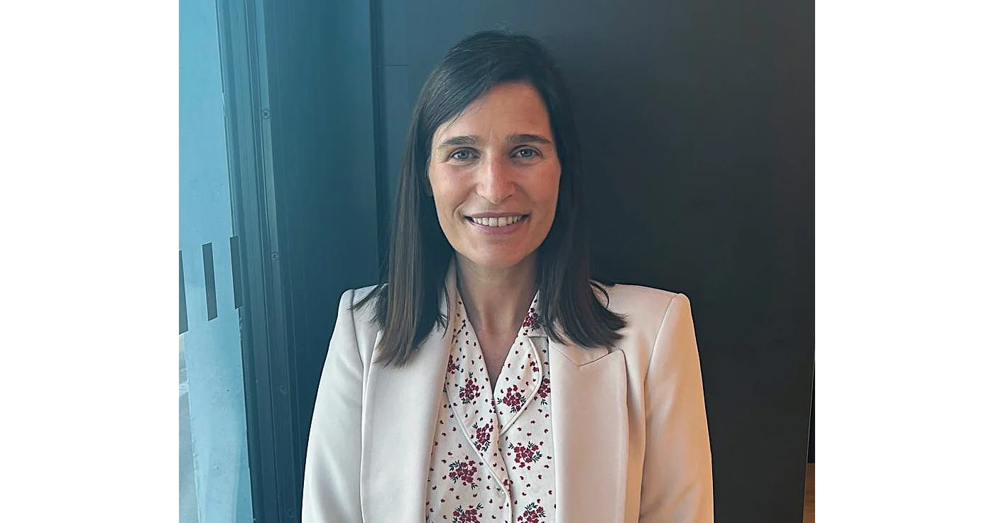 Mulher a sorrir, de blazer claro e blusa floral, junto a uma janela