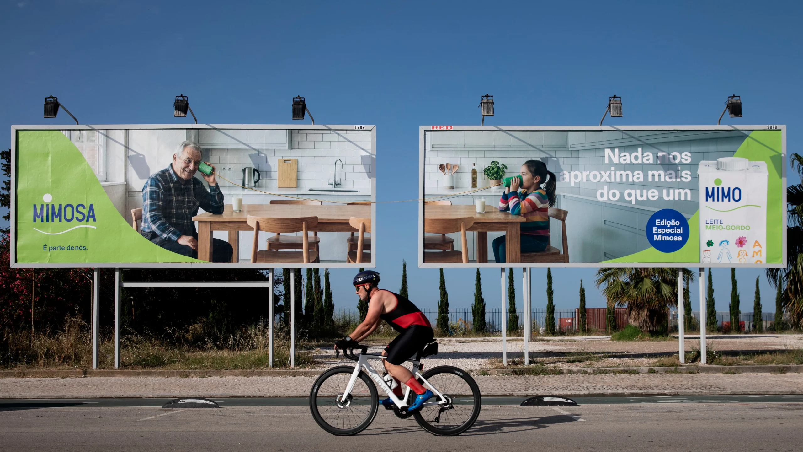 Dois outdoors da Mimosa com pessoas a beber leite por telefone, vistos por um ciclista na estrada