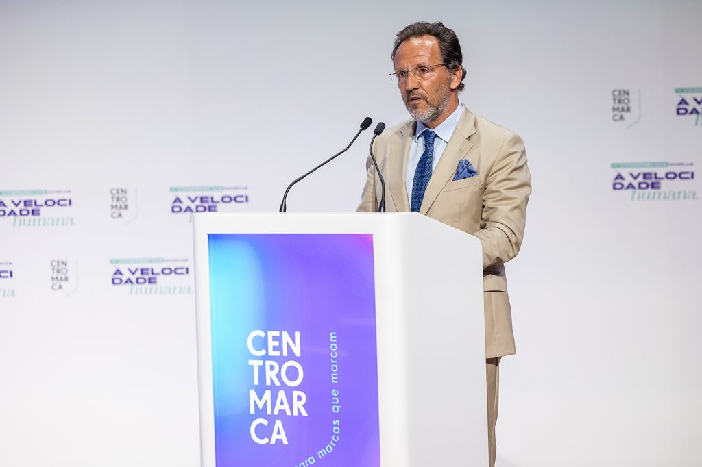 Orador em palco durante um congresso empresarial, discursando a partir de um púlpito perante uma audiência profissional. O cenário integra a identidade visual do evento, dedicado à reflexão sobre o papel das marcas, dos consumidores e da inovação num contexto de transformação económica e social.