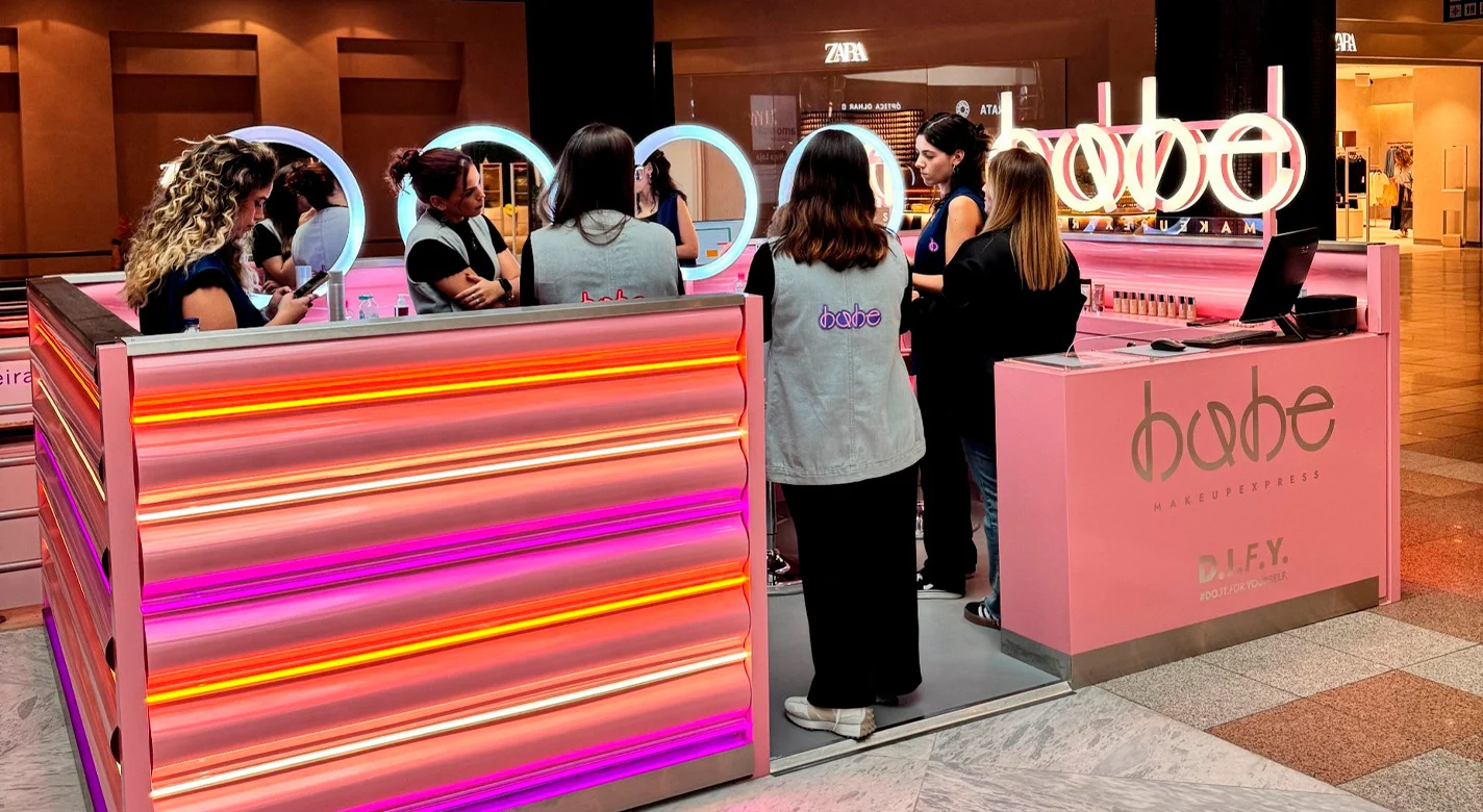 Quiosque de maquilhagem rosa num centro comercial, com várias pessoas à volta de espelhos iluminados