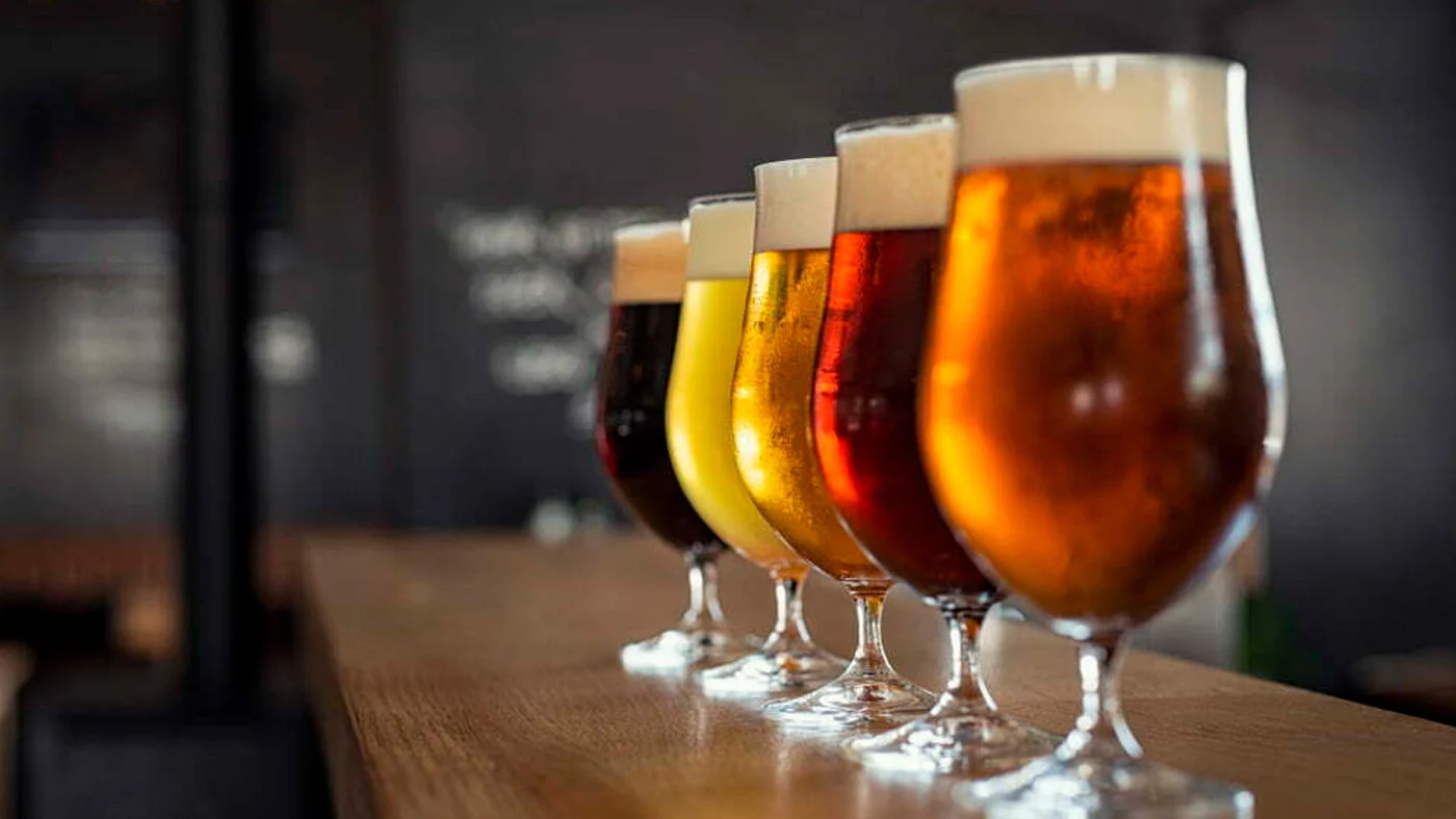 Copos alinhados com diferentes tipos de cerveja, do tom claro ao âmbar escuro, sobre um balcão de madeira
