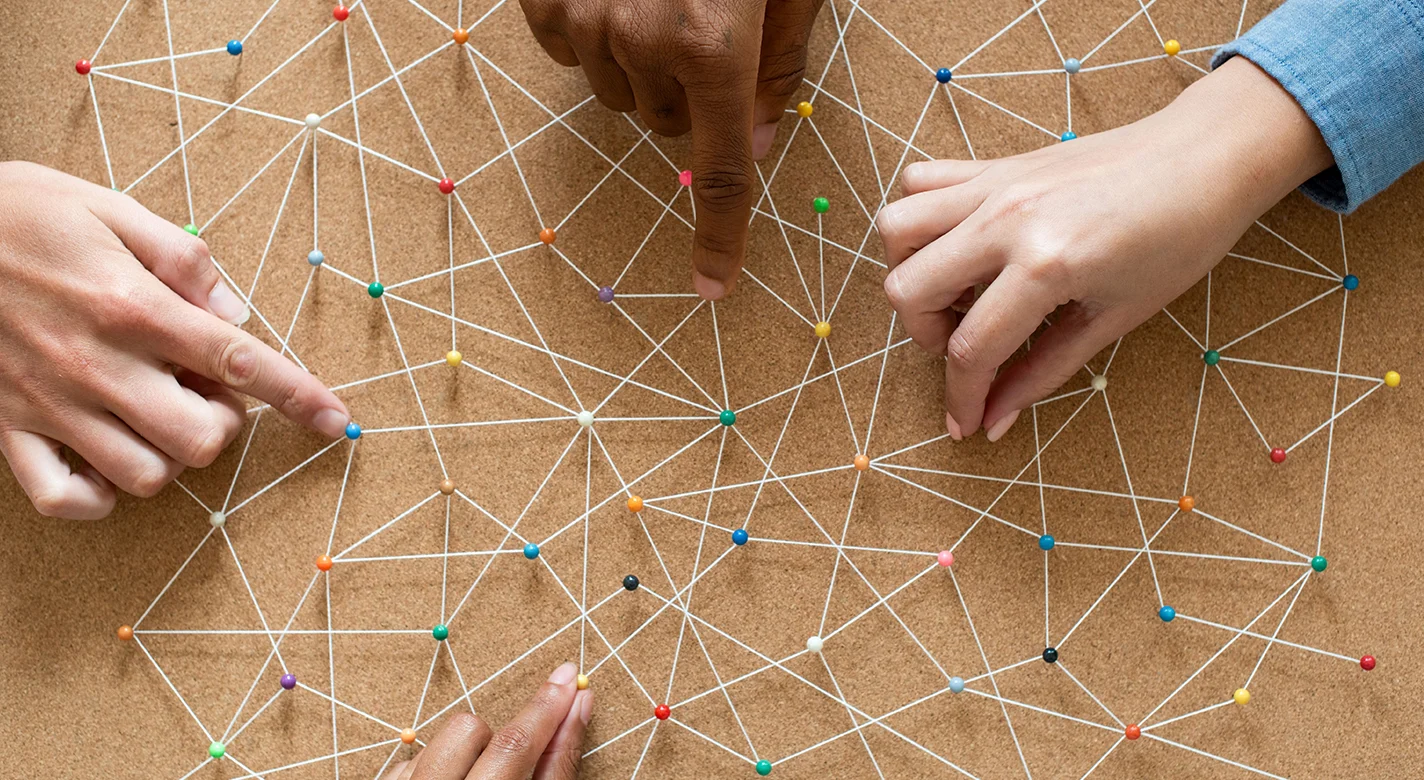 Mãos a apontar para uma rede de nós coloridos ligados por linhas brancas num quadro de cortiça