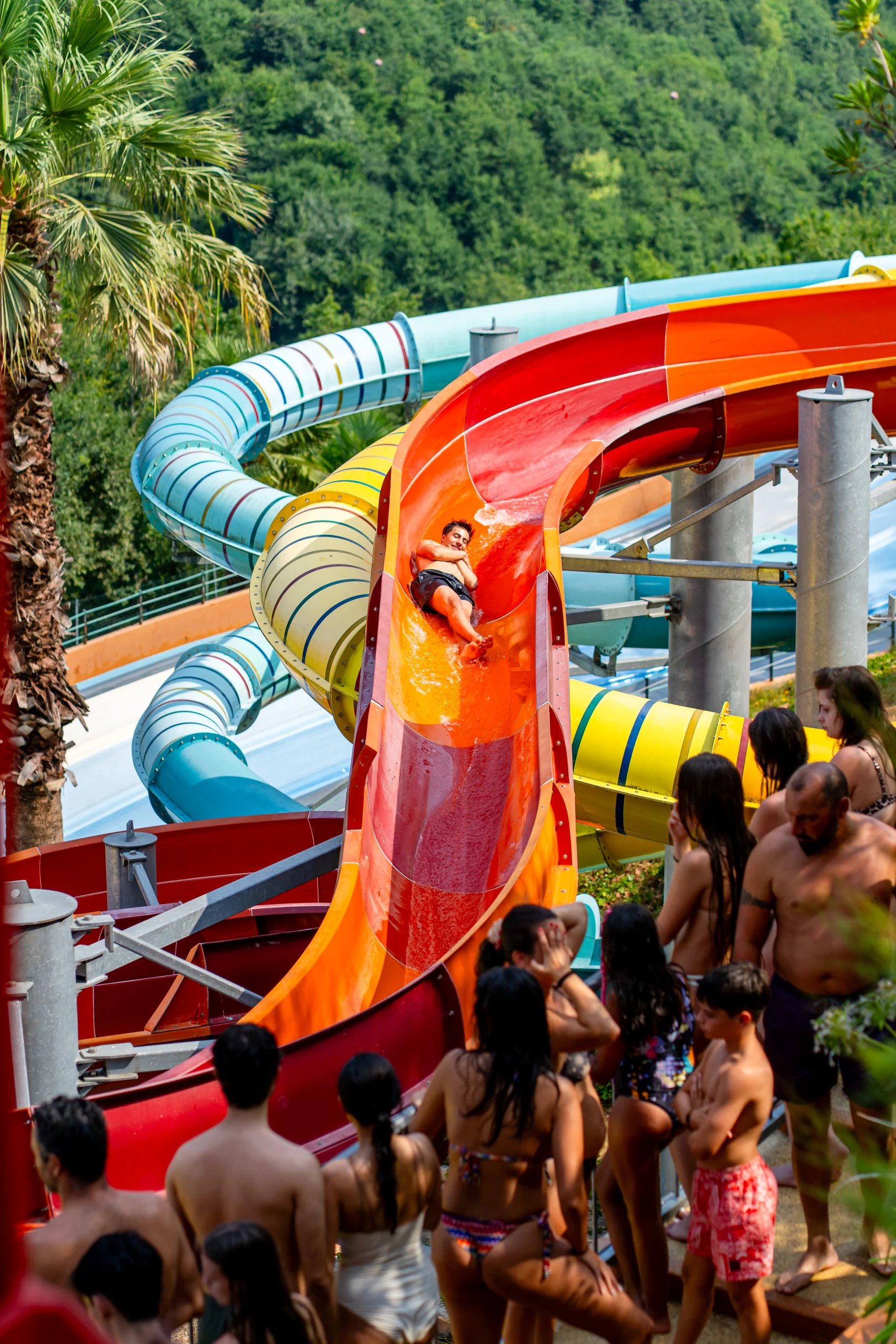 Pessoa a descer um escorrega aquático colorido num parque aquático, observada por um grupo de visitantes