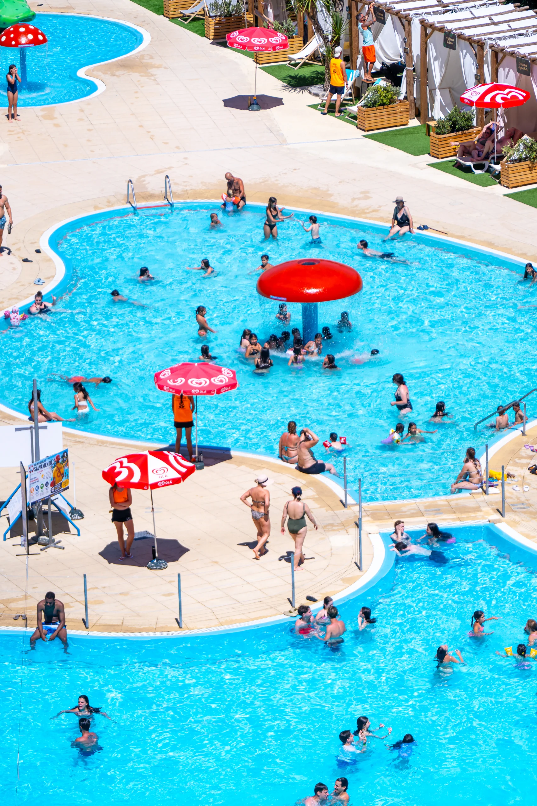 Piscina exterior cheia de pessoas a nadar e a brincar, com um cogumelo de água ao centro