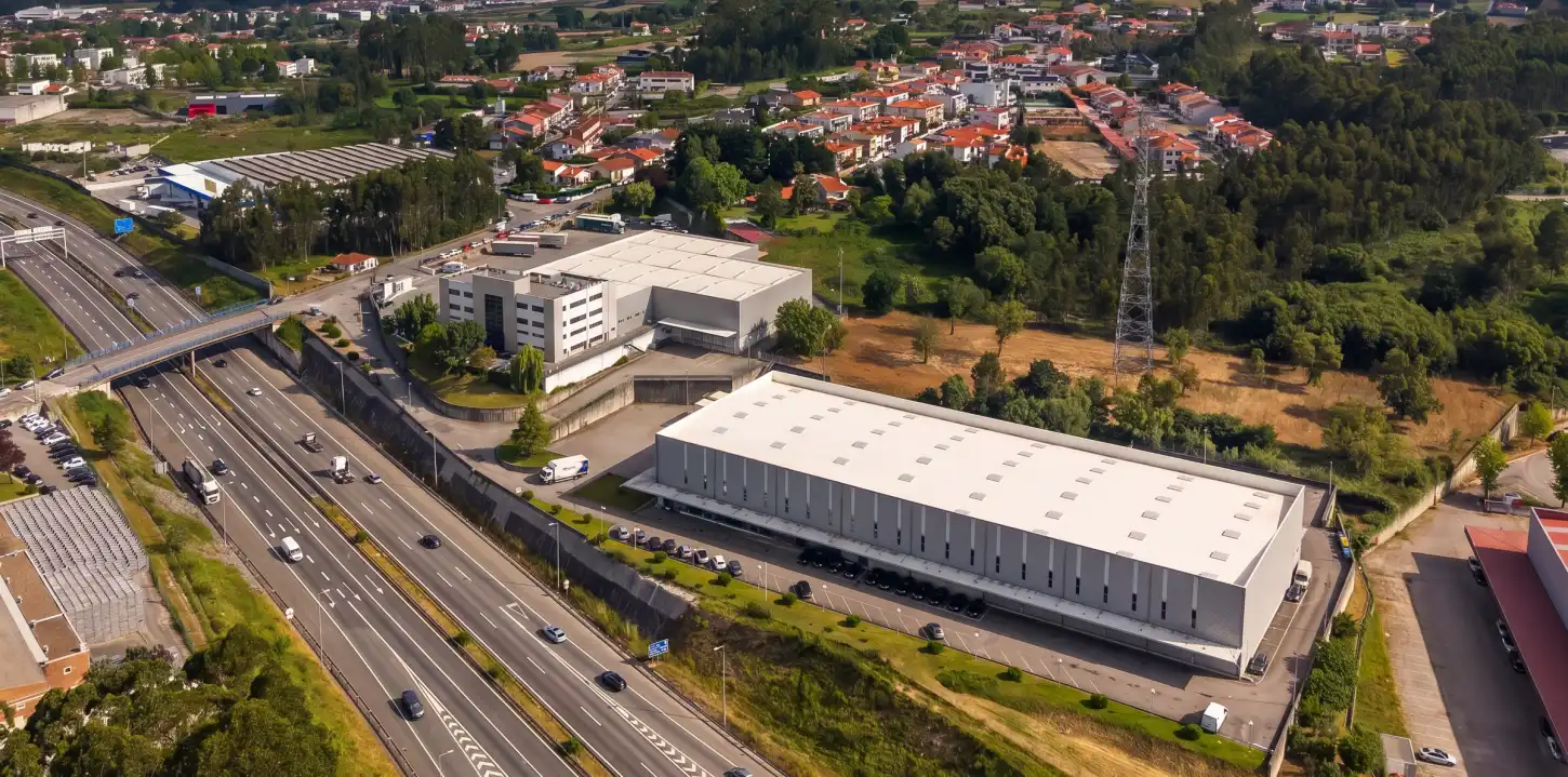 Vista aérea de uma zona industrial junto a uma autoestrada, com armazéns, casas e áreas verdes ao redor