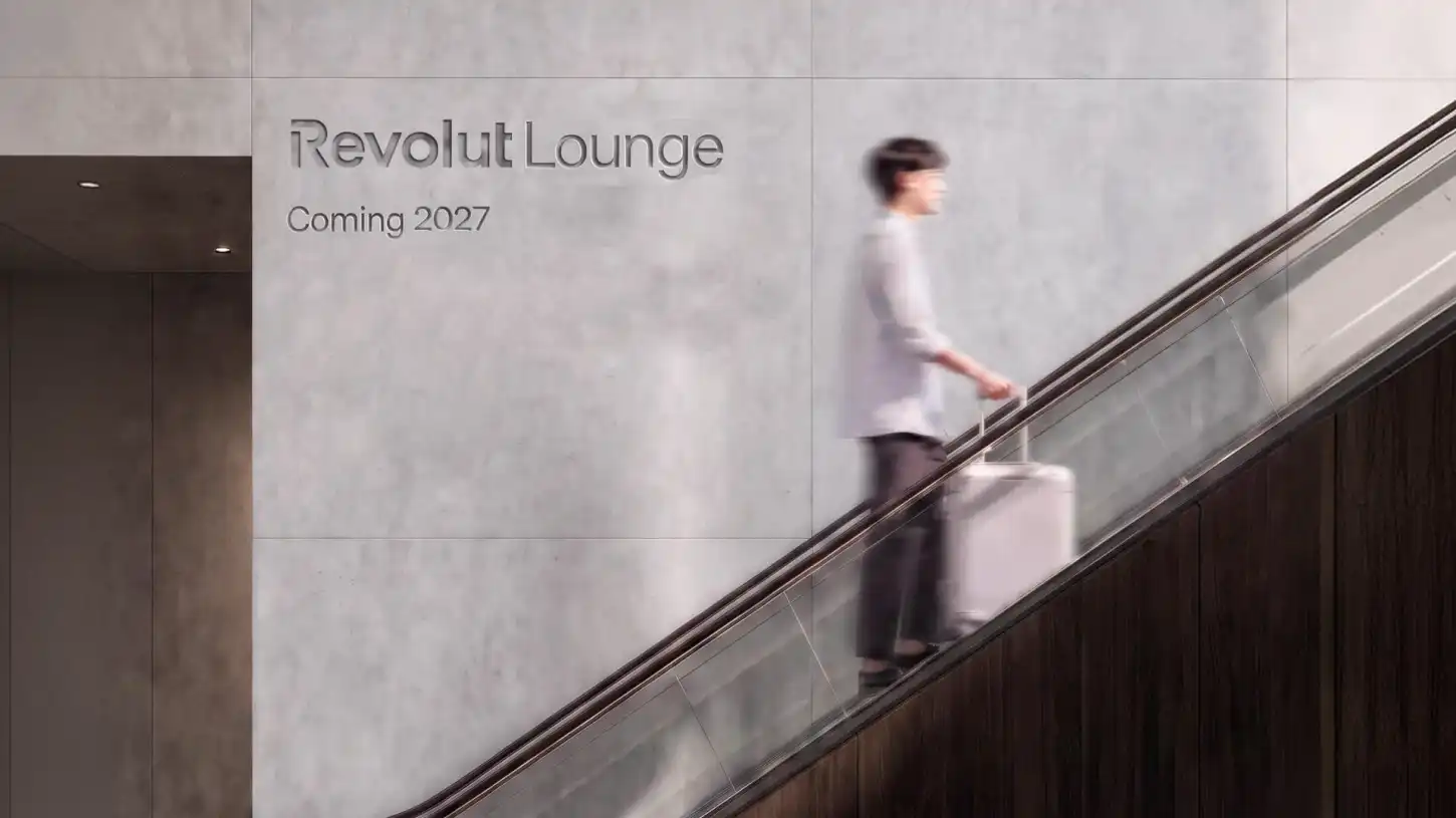 Pessoa a subir uma escada rolante com uma mala, ao lado do texto “Revolut Lounge” e “Coming 2027”