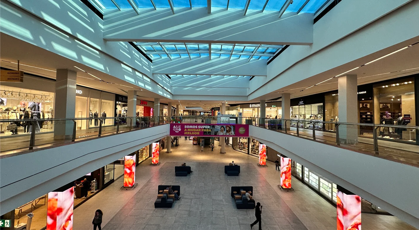 Interior de um centro comercial com lojas, claraboias, publicidade e zonas de descanso