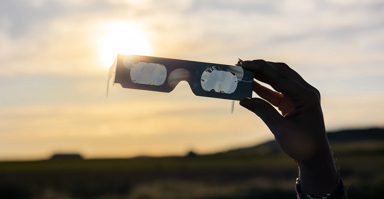 Mão a segurar óculos de eclipse contra o sol poente