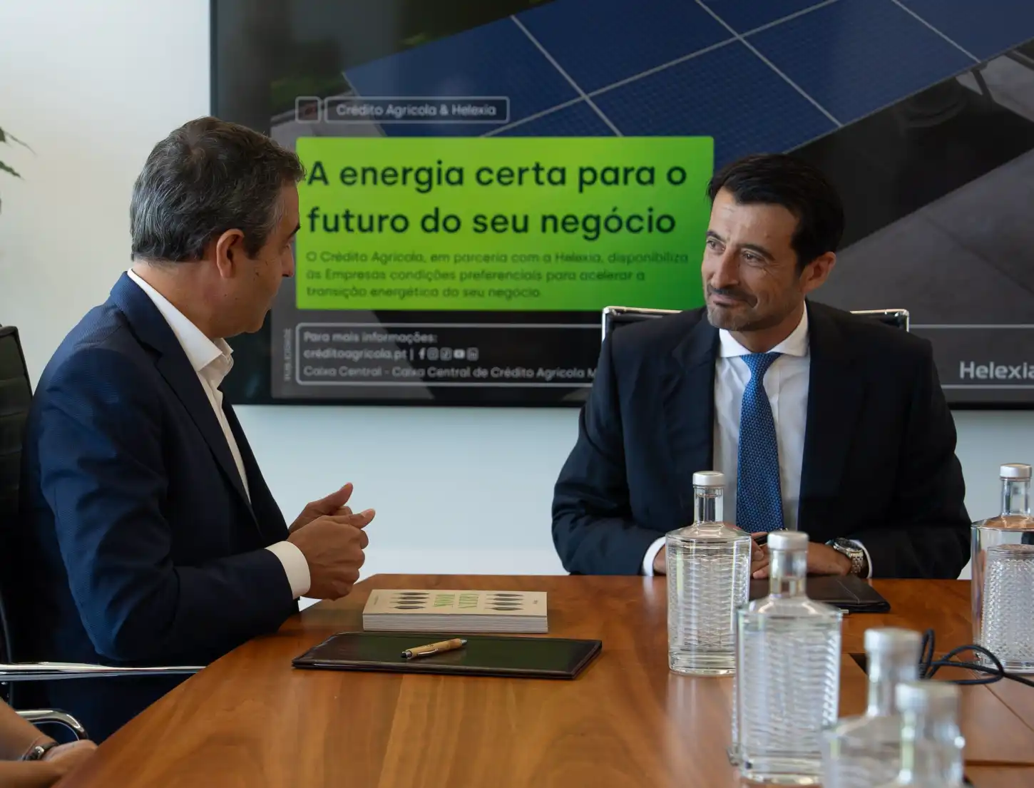 Dois homens de fato reúnem-se à mesa, com painéis solares visíveis ao fundo