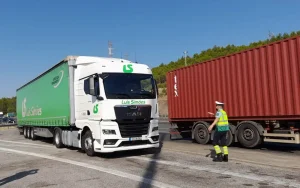 Camião de mercadorias branco e verde junto a um contentor vermelho, orientado por um agente de trânsito