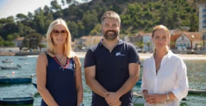 Três pessoas posam junto à água, com barcos e casas costeiras ao fundo