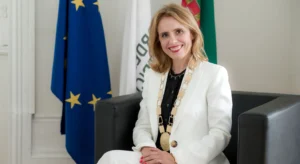 Mulher sorridente sentada numa poltrona, com as bandeiras da União Europeia e de Portugal ao fundo, usando uma cadeia cerimonial