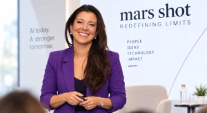 Mulher sorridente, de blazer roxo, segura um comando diante de um painel com a mensagem “mars shot: redefining limits”
