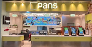 Interior de um restaurante Pans, com balcão de atendimento, quiosques de autoatendimento e ecrã com o menu