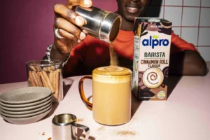 Pessoa polvilha canela sobre uma bebida quente junto a uma embalagem de bebida vegetal Alpro Barista com sabor a cinnamon roll