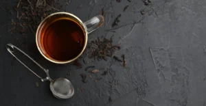 Chá âmbar numa chávena de vidro, com folhas de chá soltas e um infusor metálico sobre uma ardósia preta