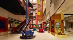 Interior de uma exposição de mobiliário moderno, com cadeiras coloridas, escadas rolantes e faixas suspensas