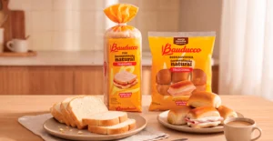 Duas embalagens de pão Bauducco, fatias de pão e sanduíches com fiambre e queijo numa mesa