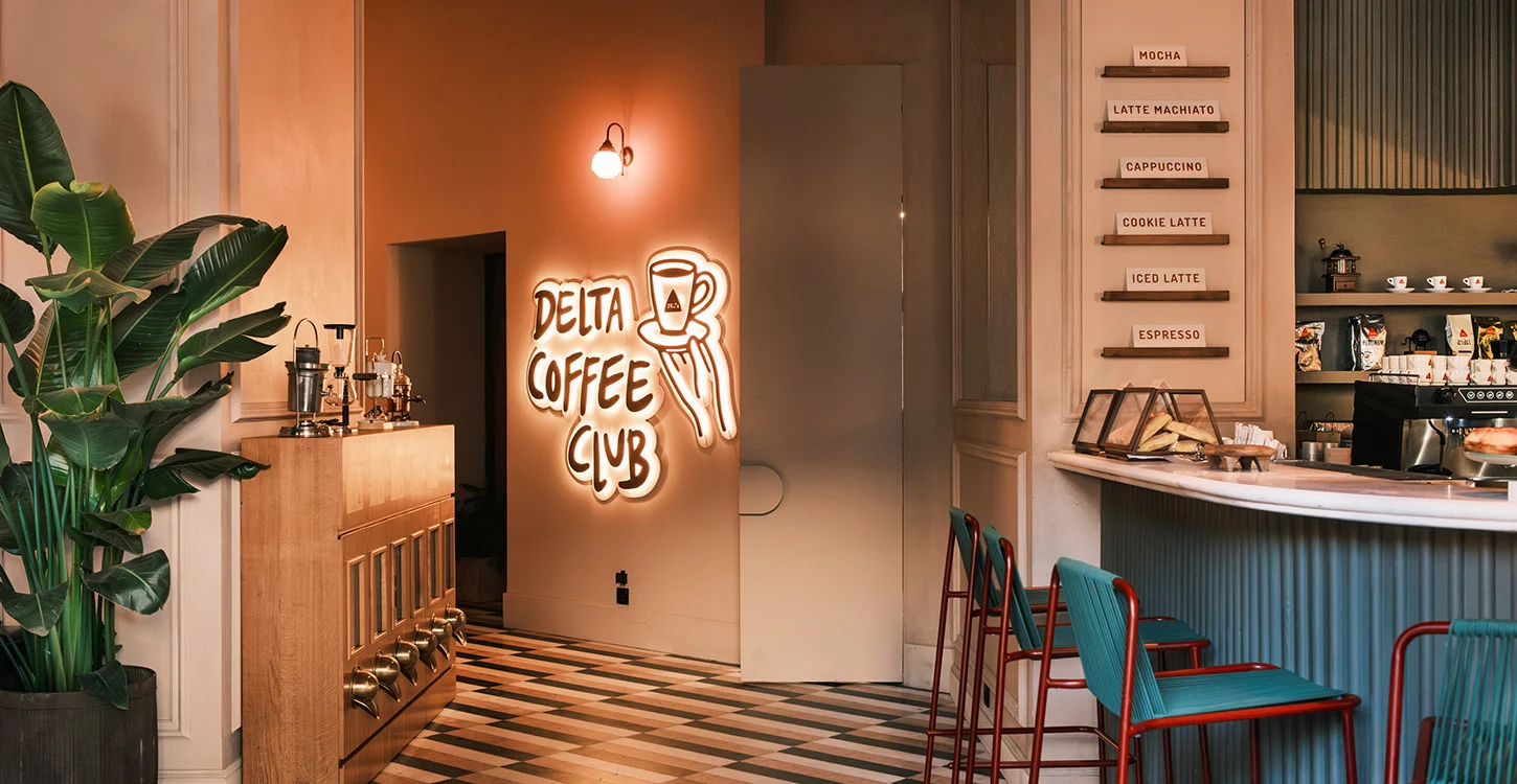 Interior de um café com balcão, equipamentos de café e letreiro luminoso «Delta Coffee Club»