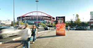 Estádio com estrutura em arco vermelho junto a uma avenida movimentada, com trânsito e peões em primeiro plano