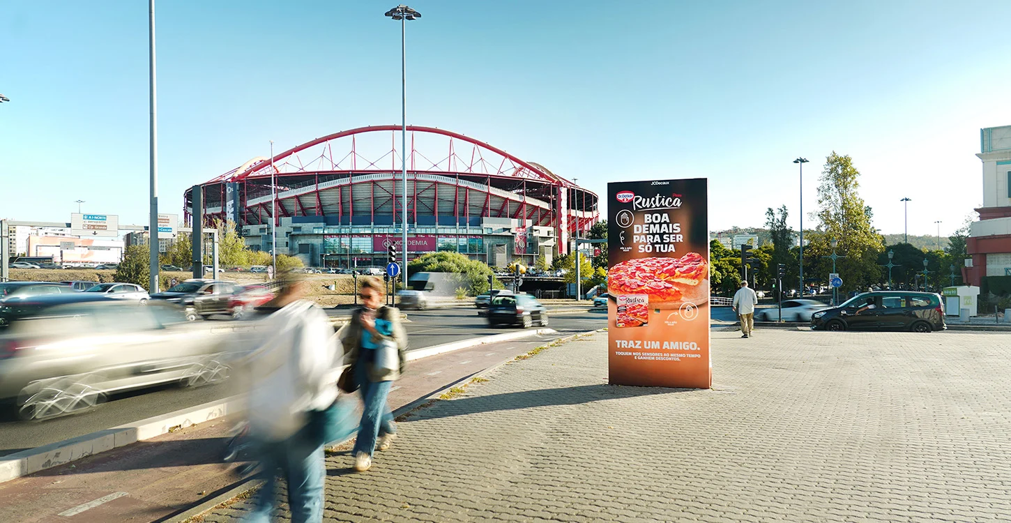 Estádio com estrutura em arco vermelho junto a uma avenida movimentada, com trânsito e peões em primeiro plano