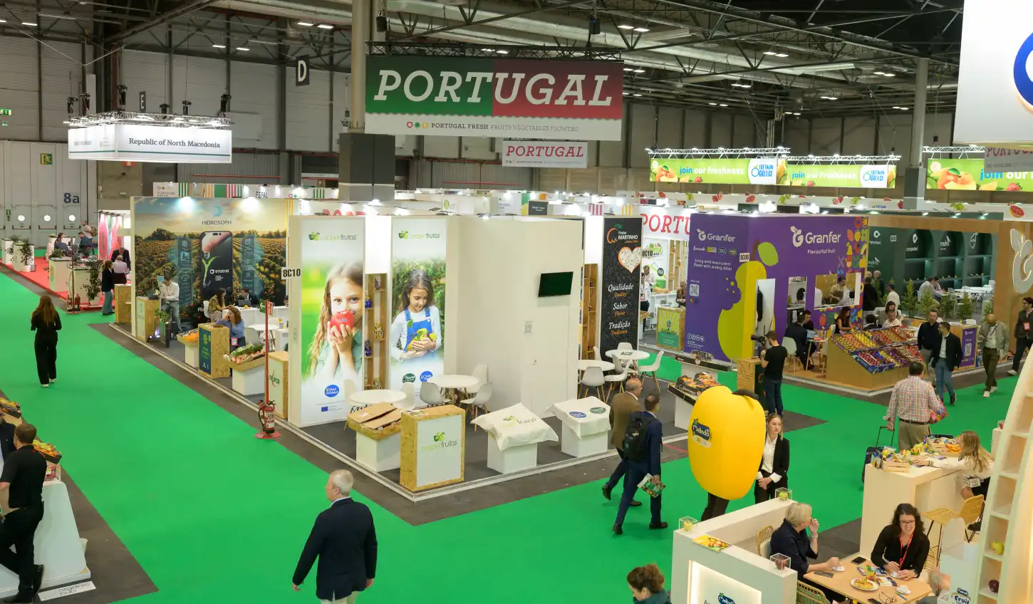 Pavilhão de uma feira comercial de frutas e legumes, com stands de empresas e visitantes, sob um grande letreiro “PORTUGAL”