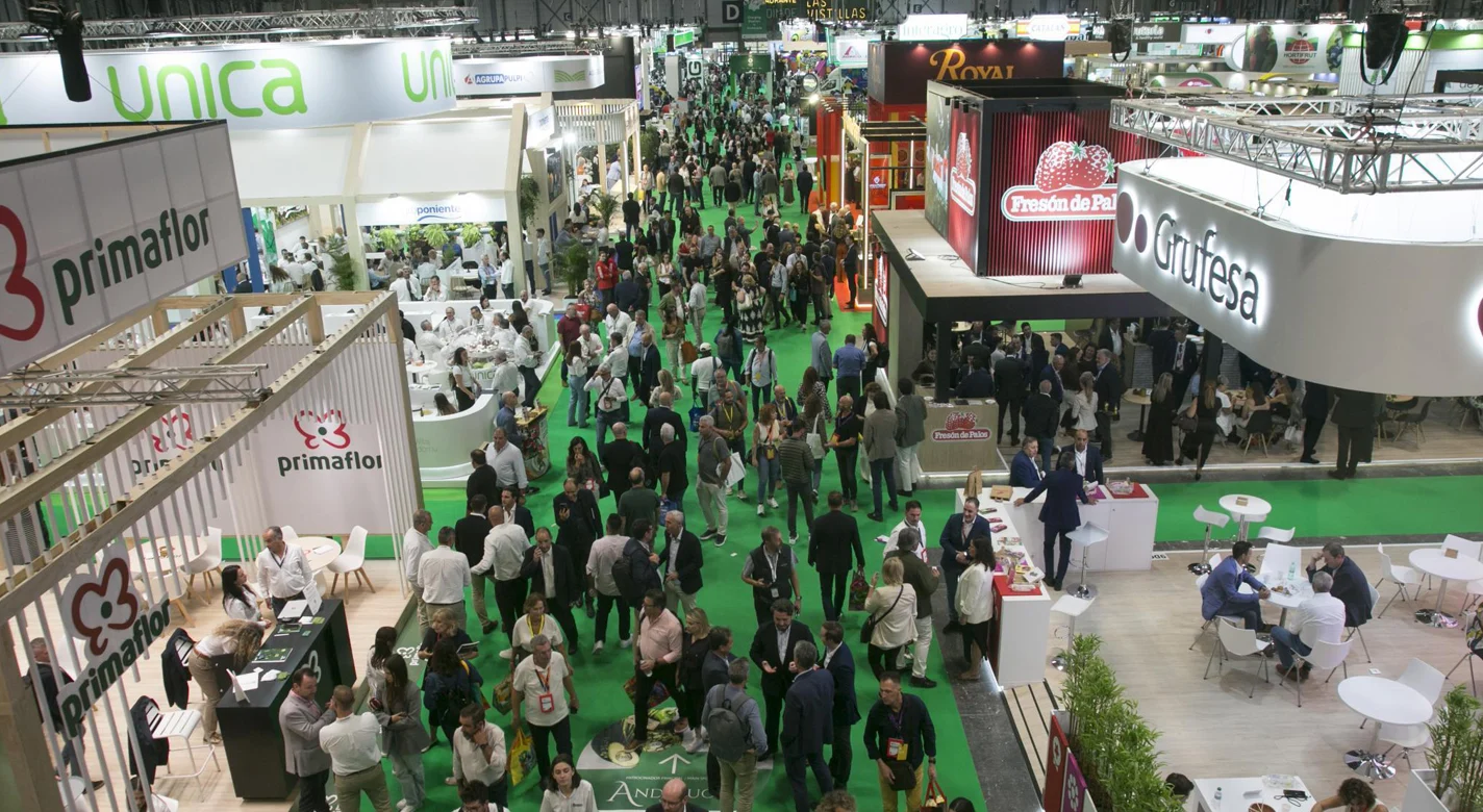 Vista panorâmica de uma feira comercial movimentada, com expositores e visitantes num pavilhão