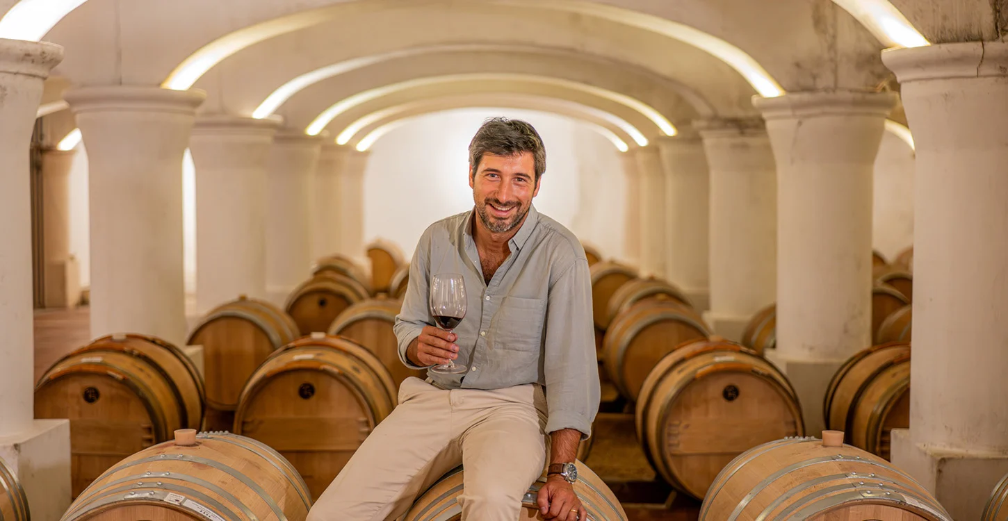 Homem sentado entre barricas de vinho, segurando um copo de vinho tinto numa adega